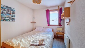 Une chambre à coucher avec un lit, une table de chevet avec une lampe, une fenêtre avec des rideaux, une étagère fixée au mur et un mur orné d’un collage de photos.