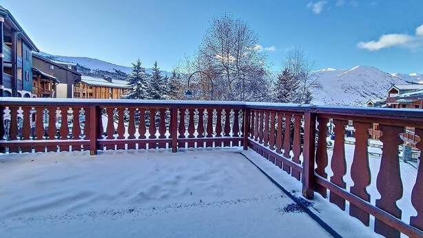 Un balcon enneigé avec une balustrade rouge, d’où l’on peut admirer un paysage montagneux et des bâtiments.