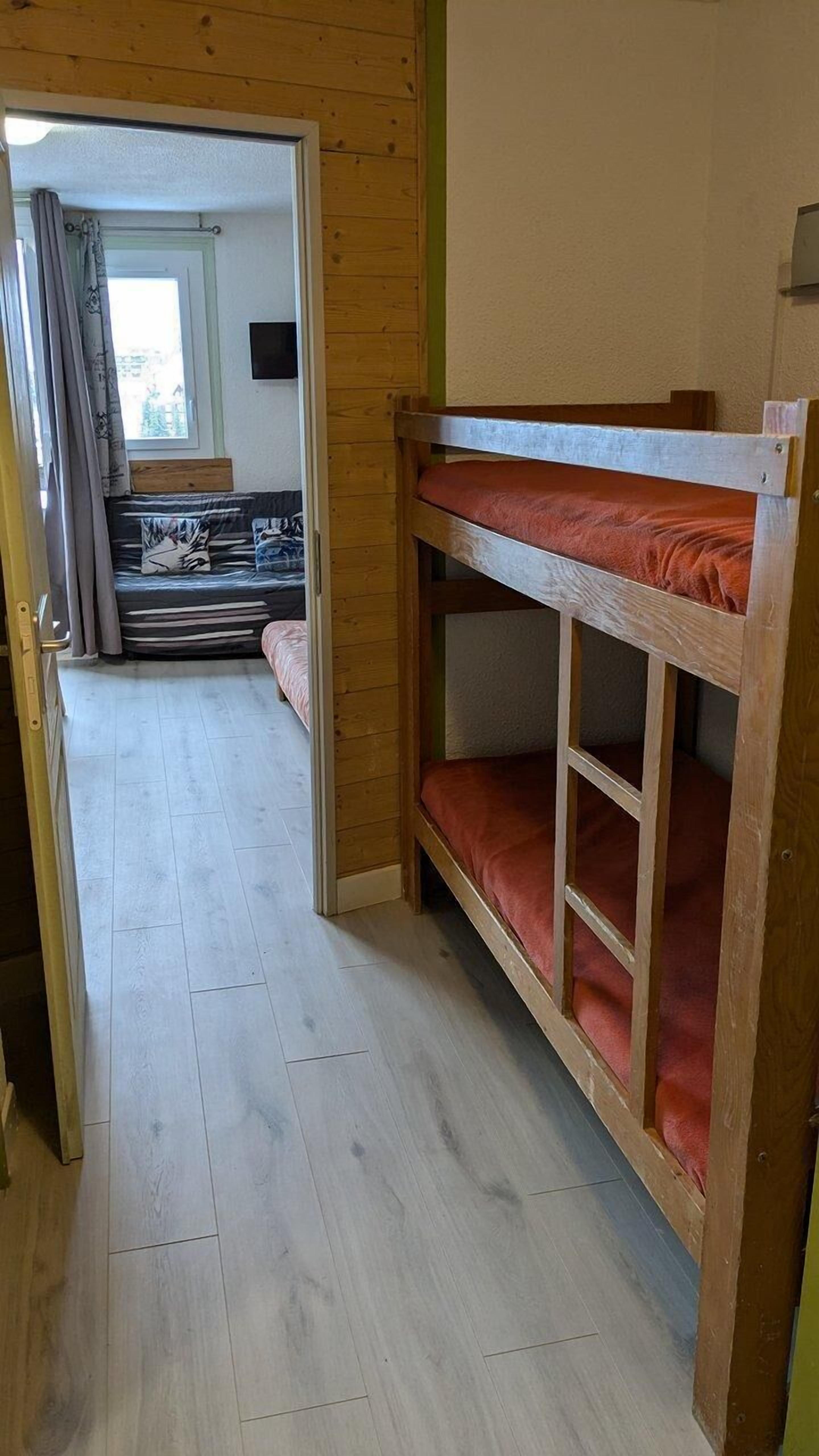 A bunk bed with a wooden frame and red bedding, a wooden floor, and a view into a room with a bed and curtains.