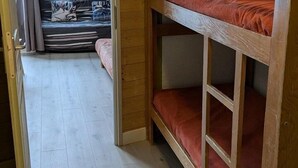 A bunk bed with a wooden frame and red bedding, a wooden floor, and a view into a room with a bed and curtains.