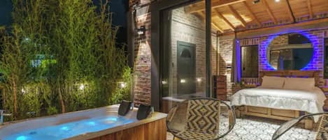 A hot tub with a wooden deck, a bed with a white duvet, and a dining area with chairs.