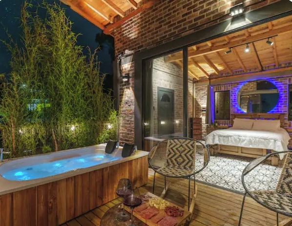 A hot tub with a wooden deck, a bed with a white duvet, and a dining area with chairs.