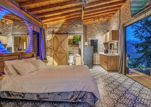 A bedroom with a large bed, wooden ceiling beams, a brick wall, and a view through a large window.