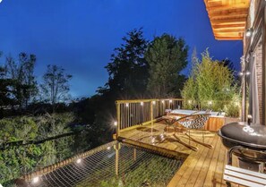 Eine Terrasse im Freien mit Grill und Sitzgelegenheit, umgeben von Bäumen und blauem Himmel.