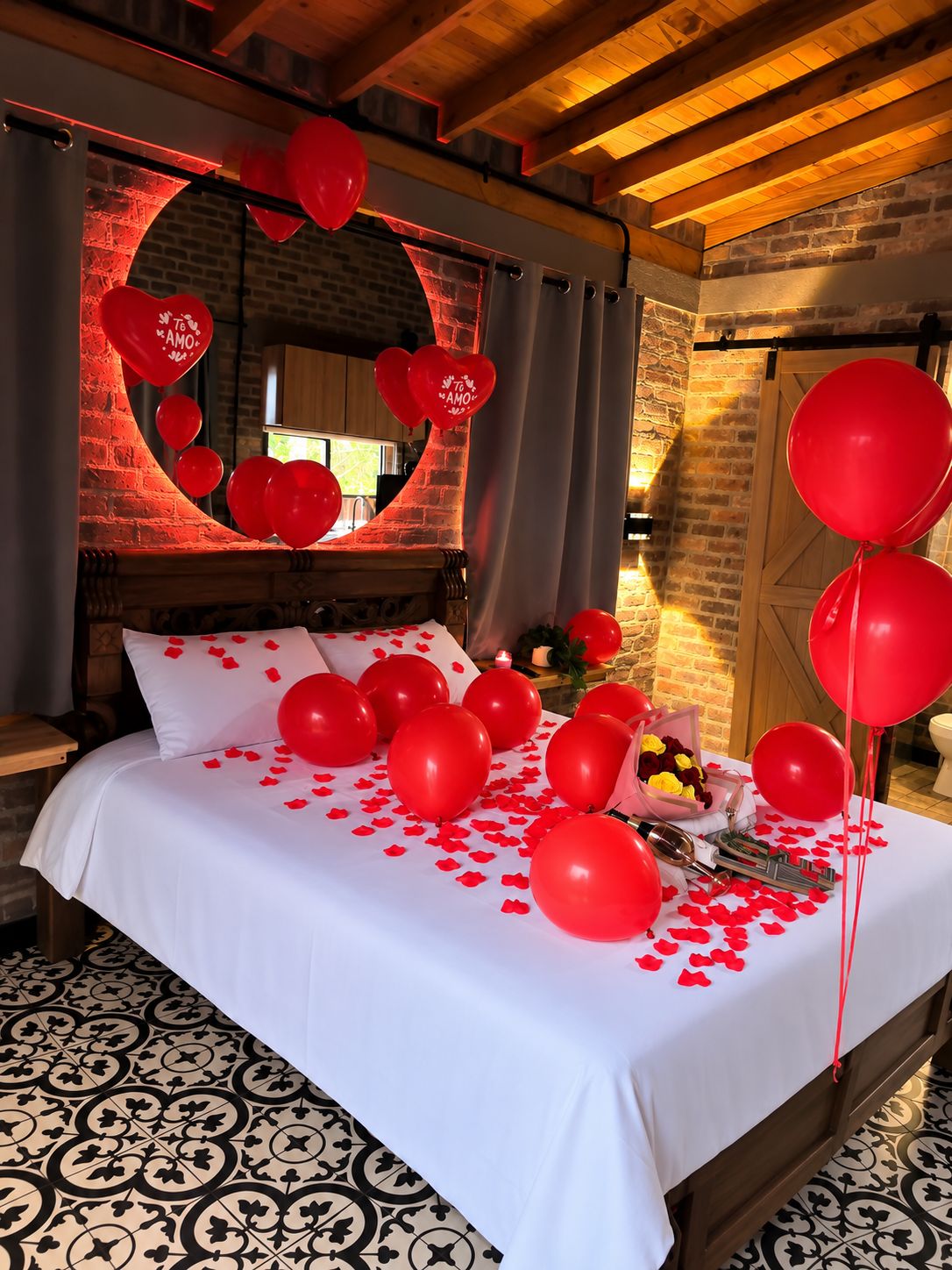 A bed with red balloons and rose petals, a brick wall, and a wooden ceiling.