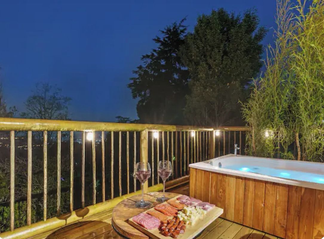 A wooden deck with a hot tub, a wooden table with a charcuterie board, and a view of the city at night.