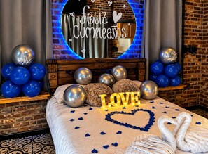 A hotel room with a bed, decorative pillows, a heart-shaped arrangement of rose petals, and a wall with a circular mirror and a message.