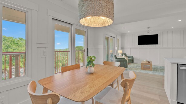 A spacious dining area with a wooden table and chairs, a hanging light fixture, and large windows offering a view of greenery.
