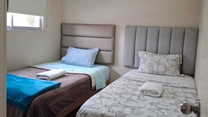 Ein Zimmer mit zwei Betten, jeweils mit Kopfstütze, einem Fenster mit Jalousie und einer Tür mit runder Klinke.