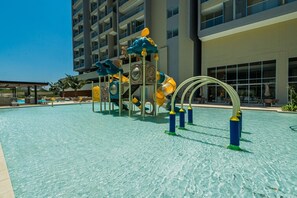 A swimming pool with a children's play structure, a covered seating area, and a modern building in the background.