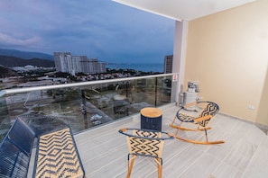 A balcony with two rocking chairs and a small table, overlooking a cityscape with buildings and a body of water.
