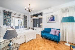 A bedroom with a bed, a blue sofa, a chandelier, a white dresser, and a window with curtains.