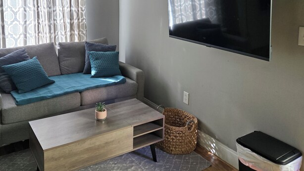 Una sala de estar con un sofá, una mesa de centro, un televisor montado en la pared y un cesto de basura.