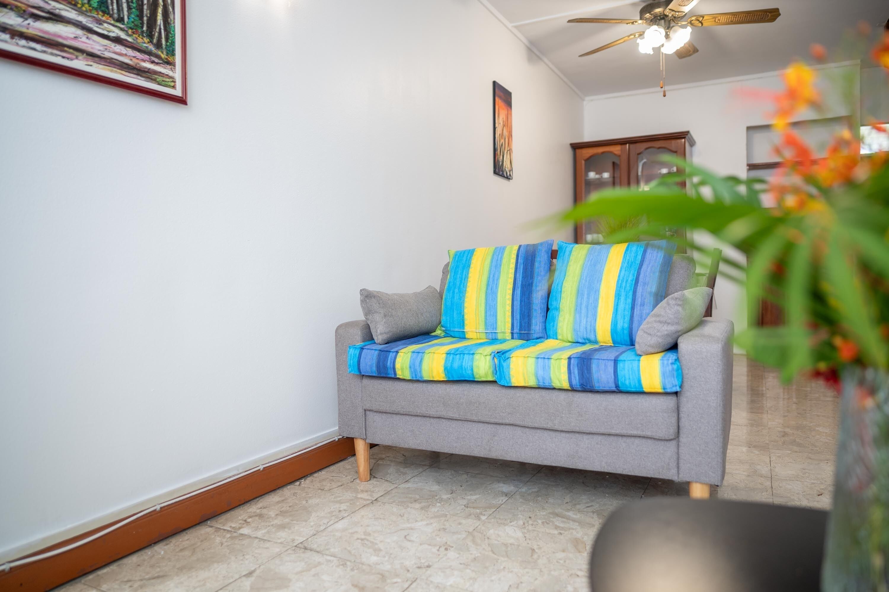 A grey sofa with blue and yellow striped cushions, a wooden cabinet, and a ceiling fan in a room with white walls and tiled flooring.