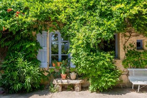 Un bâtiment rustique avec un banc en pierre, des plantes en pot et une fenêtre avec des volets.