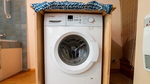 Een witte Bosch wasmachine in een kleine ruimte, met daarboven een houten plank, een betegelde vloer en een ventilatie rooster aan de rechterkant.