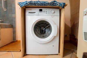 Een witte Bosch wasmachine in een kleine ruimte, met daarboven een houten plank, een betegelde vloer en een ventilatie rooster aan de rechterkant.