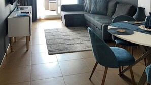 A modern living room with a sofa, dining table, and chairs. There is a TV on a stand, a window with curtains, and a ceiling light.
