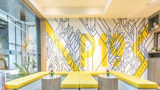 Ein modernes Interieur mit einer Wand mit geometrischem Muster, einer zentralen Sitzecke mit gelben Kissen und einem Glastrennwand links.