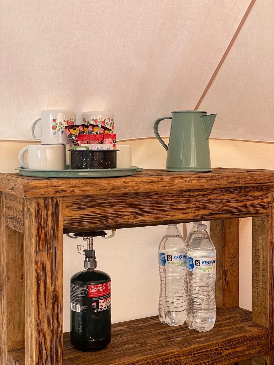 Une table en bois avec une machine à café, une tasse, un pichet et deux bouteilles d’eau.