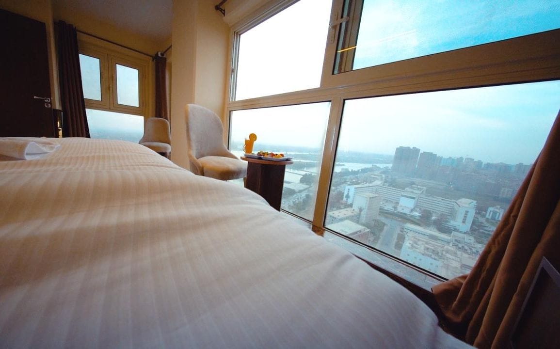 Ein Hotelzimmer mit einem großen Bett, einem Sessel, einem kleinen Tisch mit einer Speisenplatte und Blick auf eine Stadt durch große Fenster.