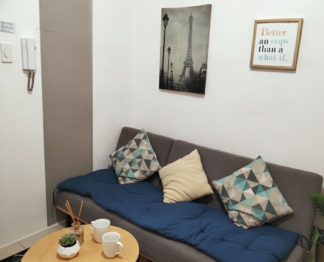 Una habitación con un sofá, dos cojines, una mesita con una planta y dos tazas, un teléfono en la pared, un cuadro con la Torre Eiffel y una cita enmarcada.