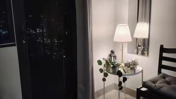 Un dormitorio moderno con un ventanal grande, una mesita de noche con lámpara y vistas a la ciudad de noche.