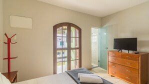 Habitación de hotel con cama, televisión, cómoda de madera, perchero y puerta de vidrio que da al exterior.