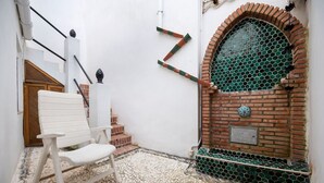Uma cadeira branca em um pátio com piso de pedrinhas, uma parede branca com uma porta de madeira, uma parede de tijolos com uma janela em mosaico verde e uma placa de metal.