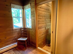 A wooden cabin interior with a wooden chair near a window, a door leading outside, and a potted plant.