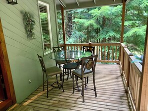 A wooden deck with a glass table and chairs, surrounded by a railing, overlooking a forested area.