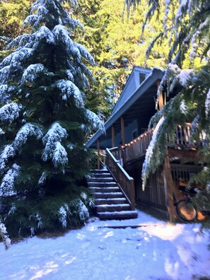 Eine Holzhütte mit einer Treppe zu einer Holzterrasse, umgeben von schneebedeckten Bäumen.