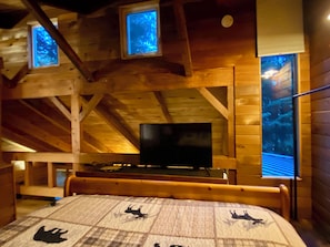 Une cabane en bois avec un lit, un téléviseur et deux fenêtres éclairées de lumière bleue.