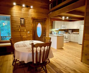 A wooden cabin interior with a dining table, chairs, a kitchen area, and a large window.