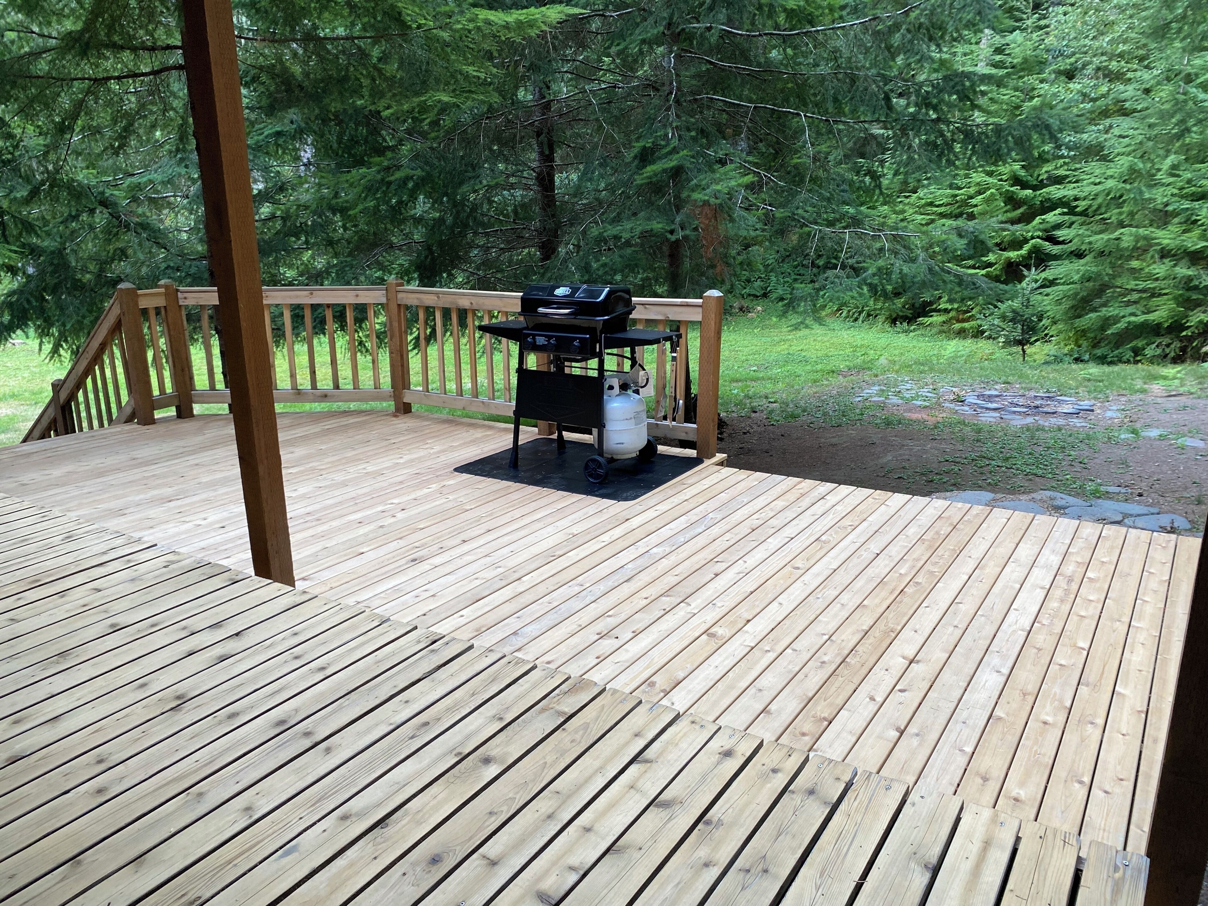A wooden deck with a grill and propane tank, surrounded by a wooden railing and dense forest.