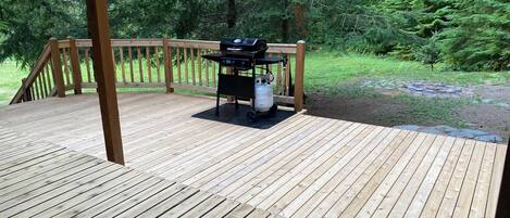 A wooden deck with a grill and propane tank, surrounded by a wooden railing and dense forest.