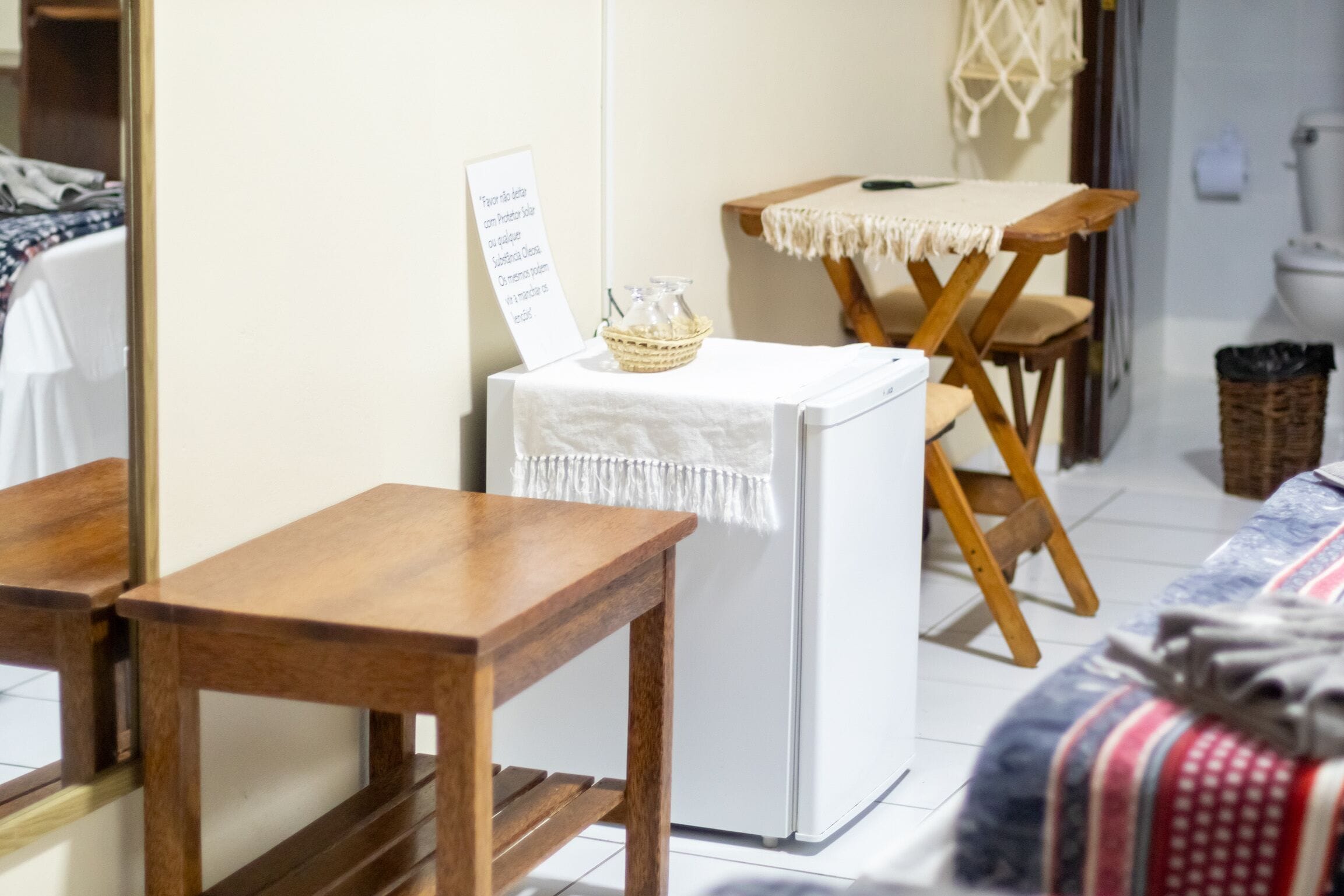 A small kitchenette with a refrigerator, a wooden table, and a chair. There is a sign on the refrigerator and a basket on the table.