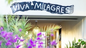 Une enseigne portant l’inscription « VIVA MILAGRES » est placée au-dessus de l’entrée d’un immeuble, et on peut y voir des plantes en pot et un luminaire suspendu.
