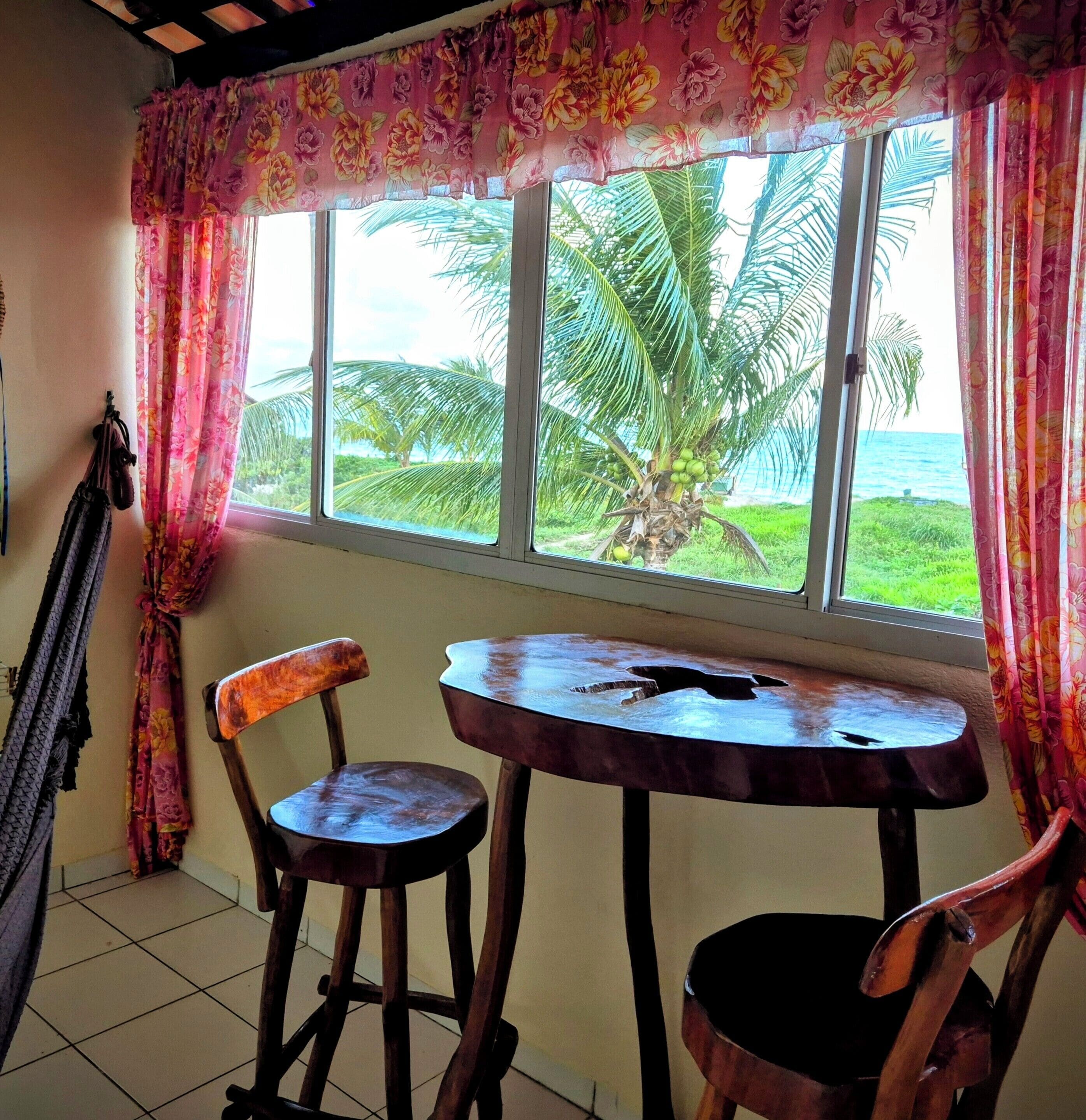 A room with a window overlooking a palm tree and ocean view, a wooden chair, a small round table, and a hammock.