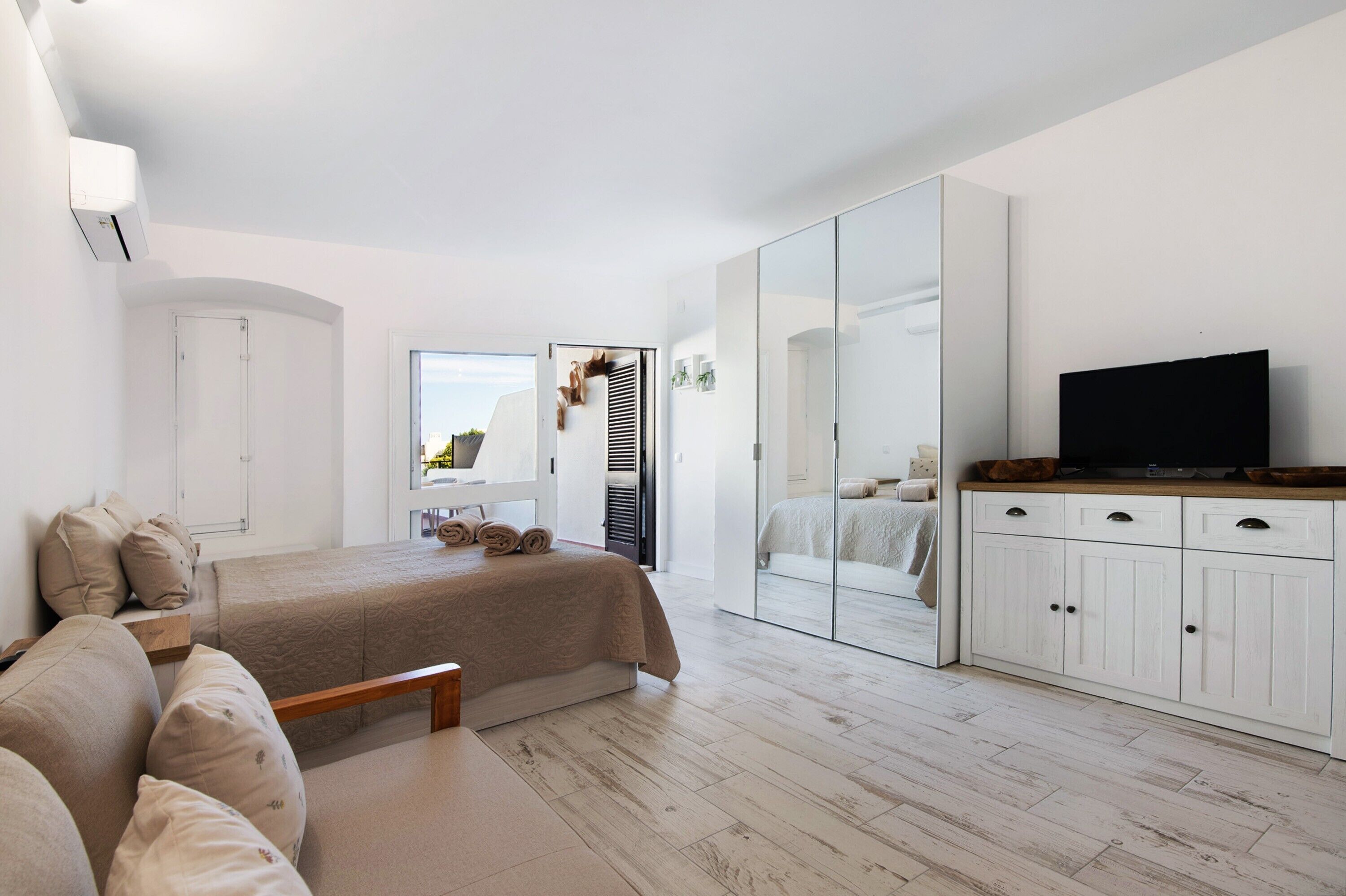 Quarto com cama, sofá, televisão, guarda-roupa e porta de correr para uma varanda.