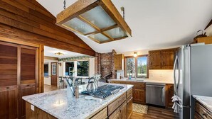 A kitchen with wooden cabinetry, a large island, a brick fireplace, and a stainless steel refrigerator. - Swan River Rendezvous - Riverfront Retreat w/Dock (Bigfork)