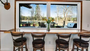 A bar area with three wooden chairs, a large window with a view of outdoor seating, and a mounted deer head. - Swan River Rendezvous - Riverfront Retreat w/Dock (Bigfork)