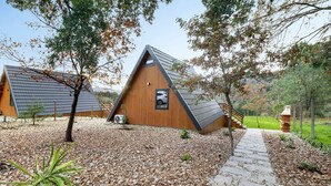 Une cabane triangulaire en bois, accessible par un sentier de pierres, entourée d’arbres et de gravier.