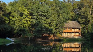 Una cabaña junto al lago con un porche y un espacio cubierto, rodeada de árboles frondosos.