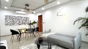 Una sala moderna con un sofá gris, un sillón amarillo, un comedor con mesa y sillas, y una gran obra de arte abstracta en la pared.
