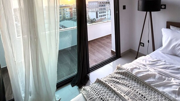 Una habitación de hotel con cama, un ventanal con cortinas y vistas a la ciudad, además de una lámpara de pie.