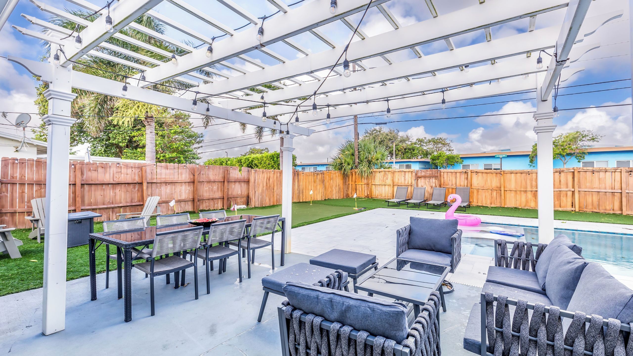 A covered outdoor patio with a pool, lounge chairs, and a wooden fence in the background.