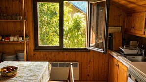 Een houten keukentje met een eettafel, een raam met uitzicht op groen en eenvoudige keukenvoorzieningen.