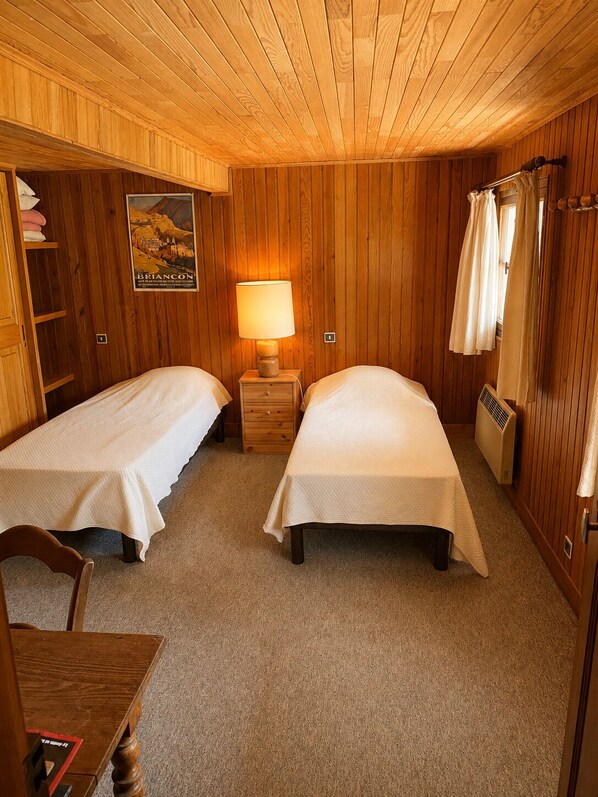Une chambre avec deux lits simples, un plafond en bois, une affiche au mur, une table de chevet avec une lampe et un radiateur.