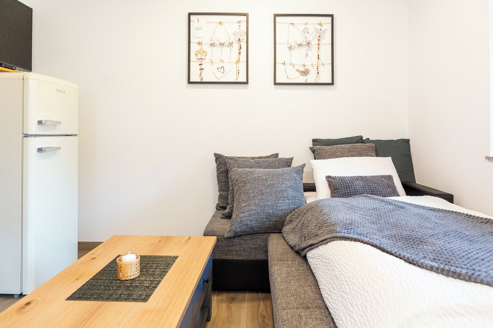 Ein kleiner, moderner Raum mit einem Bett, einem Schreibtisch und einem Kühlschrank. An der Wand hängen zwei gerahmte Kunstwerke.